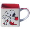 Caneca quadrada branca com interior vermelho, estampa do Snoopy segurando uma xicara de caf e um donut, sentada em cima de um cobertor vermelho.