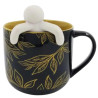 Caneca de cermica preta com detalhes dourados de folhas e interior amarelo, acompanhada de um mini boneco branco que se encaixa na borda, proporcionando um toque relaxante e divertido ao momento do ch.